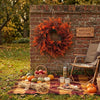 An artificial eucalyptus wreath made of orange and red foliage hangs on a brick wall surrounded by autumn harvest decor.