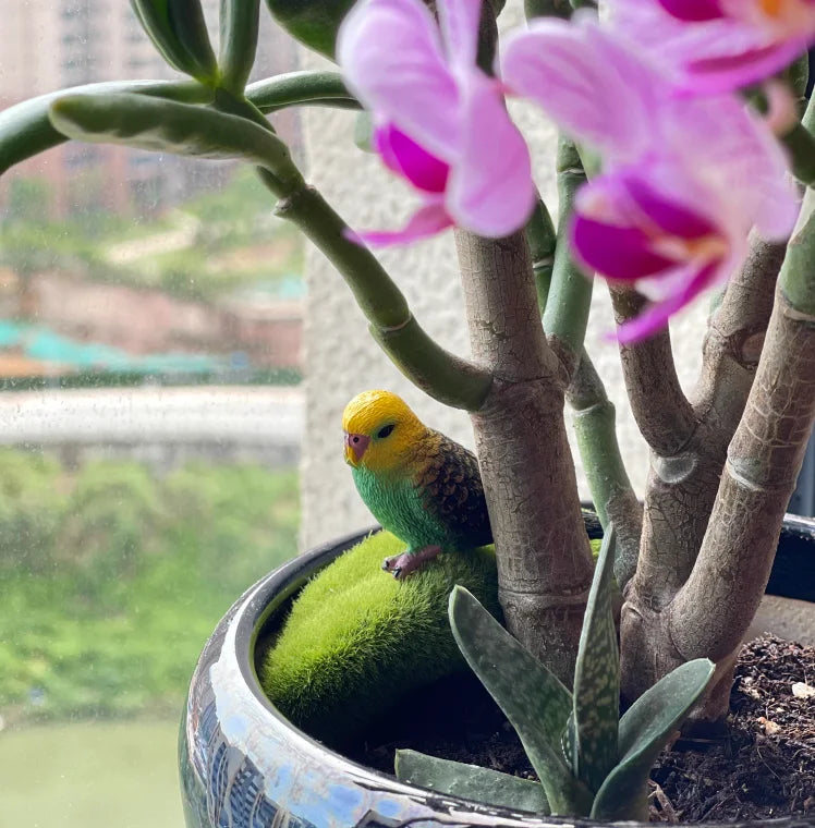 A yellow parrot figurine is perched on a plant branch in a potted indoor garden setting.