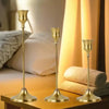 A set of three matte gold taper candle holders displayed on a wooden surface next to bedding.