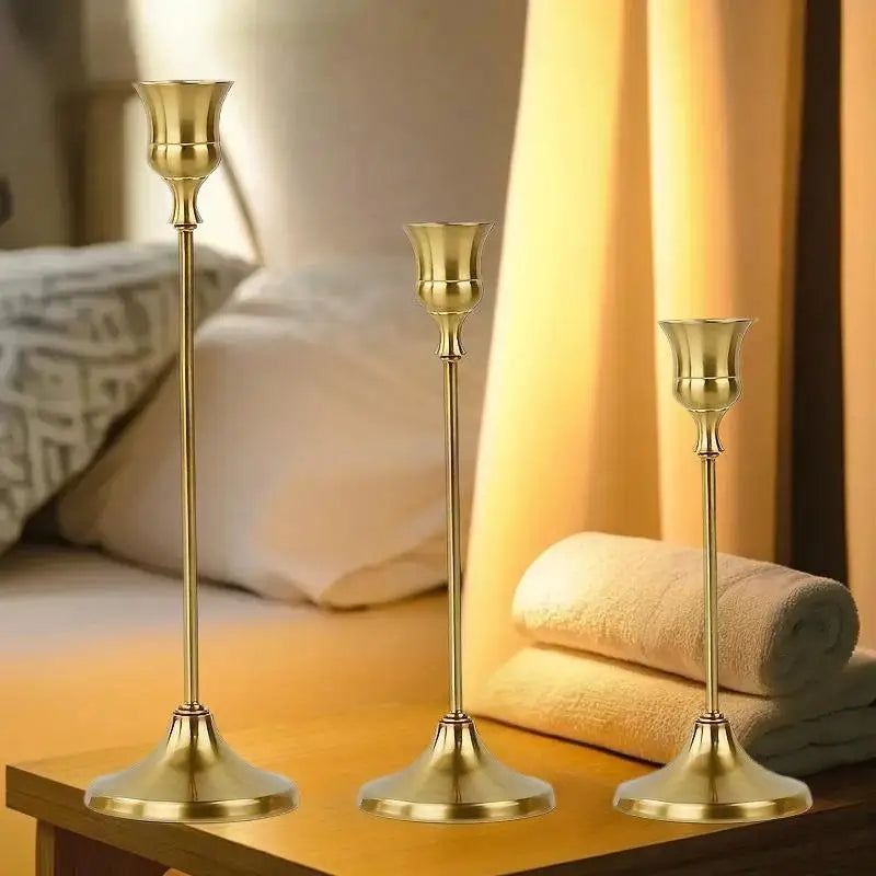 A set of three matte gold taper candle holders displayed on a wooden surface next to bedding.