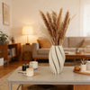 A tall vase with vertical textured waves in soft beige tones sits on a light gray table.