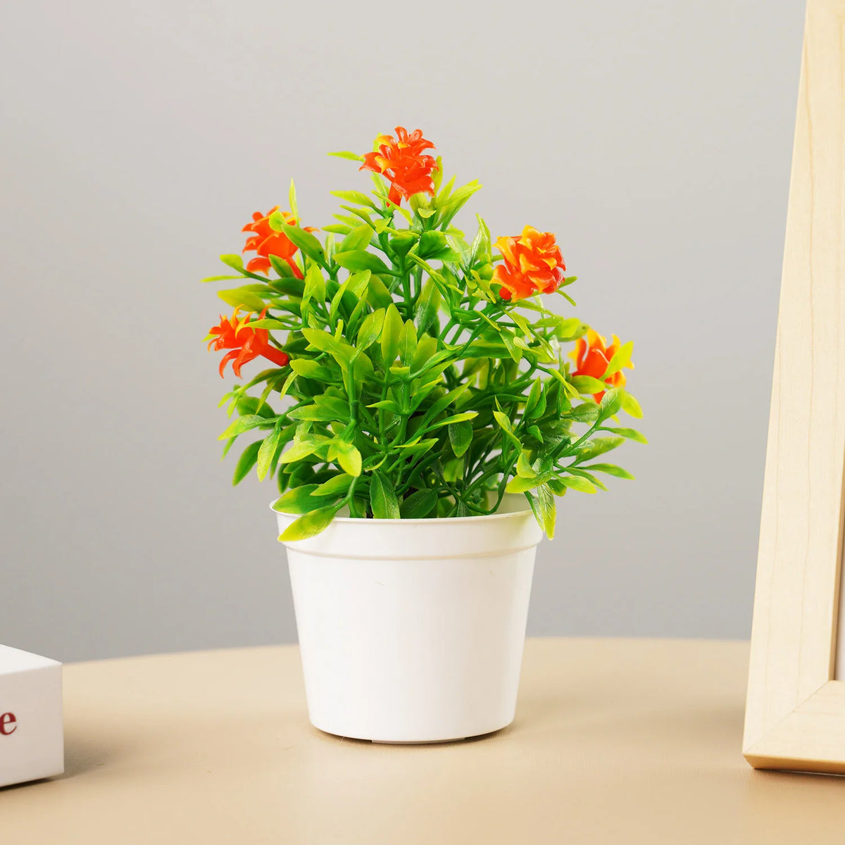 A potted artificial bonsai tree with bright orange flowers and vibrant green leaves on a light background.