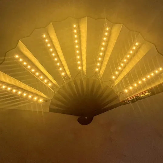 An illuminated folding hand fan with LED lights is displayed, showing a bright yellow light radiating from the fan's structur