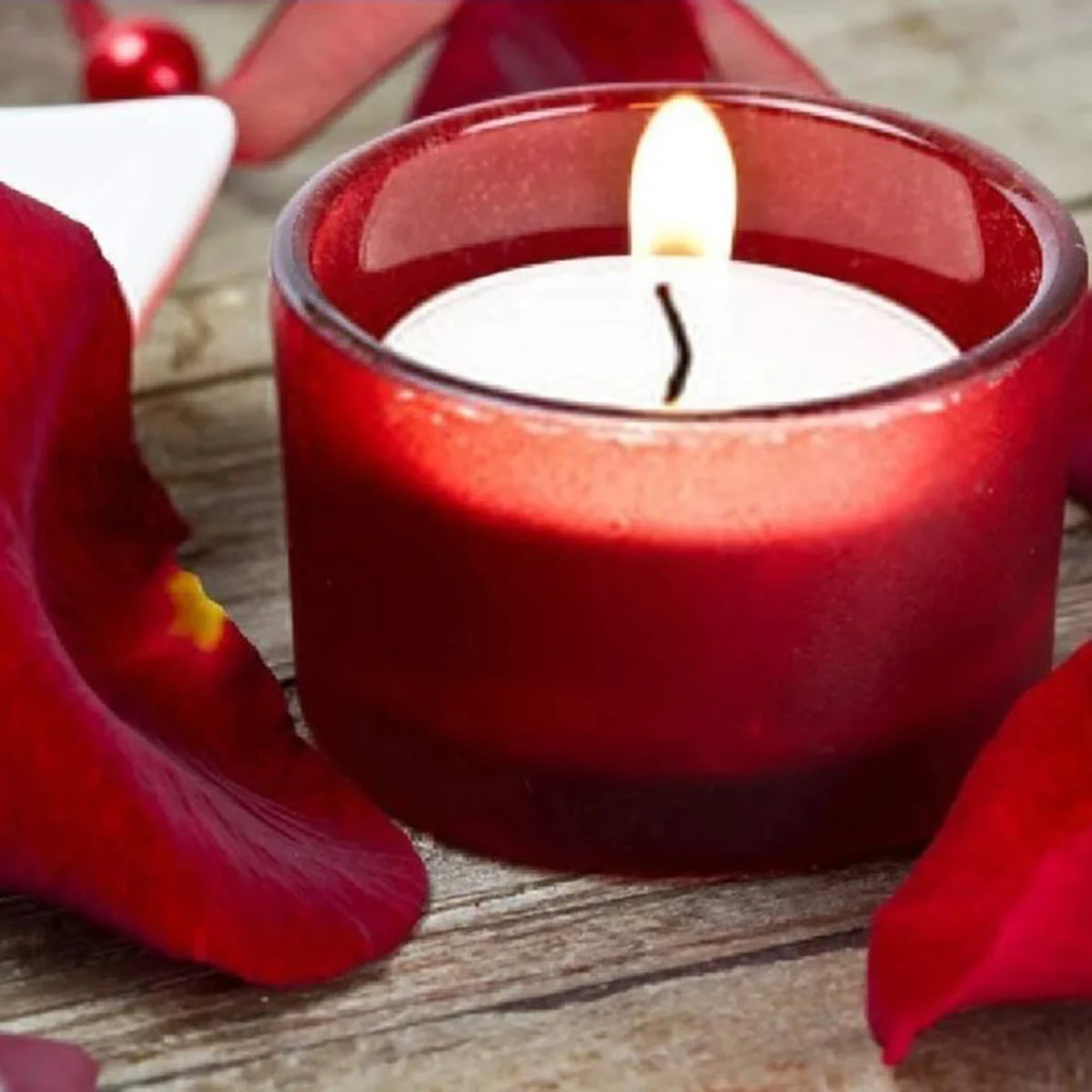 A red candle in a glass holder with a lit wick, surrounded by red rose petals on a wooden surface.