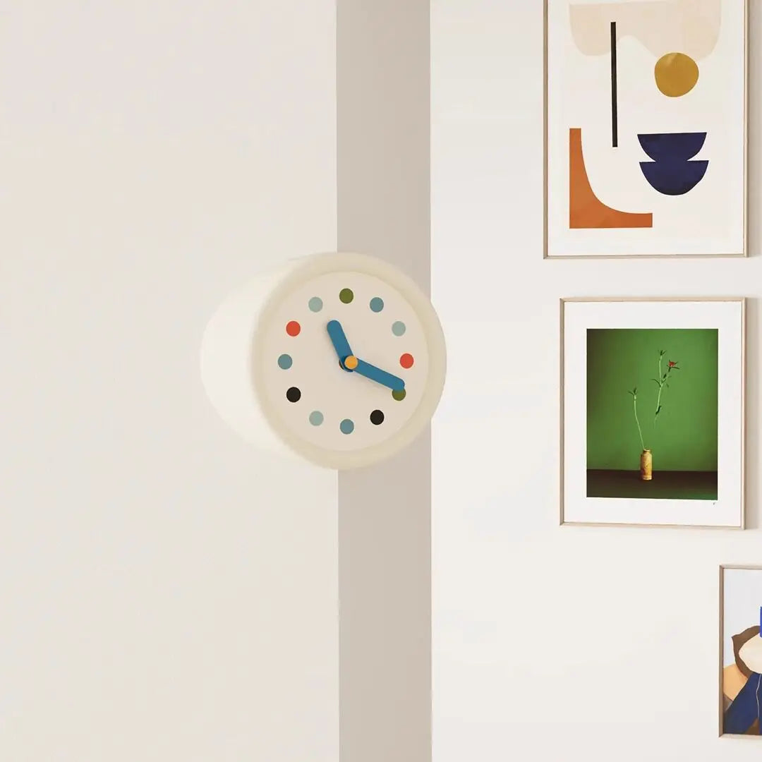 A modern resin wall clock with a light beige face and blue hands is displayed on a white wall next to framed artwork.
