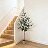 A tall artificial olive tree in a pot stands next to a modern staircase with illuminated steps.