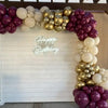 A balloon garland arch featuring burgundy, gold, and white balloons decorating a white wooden backdrop.