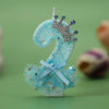 A light blue glitter candle shaped like the number 2 with a silver crown decoration and a sheer blue tulle skirt.