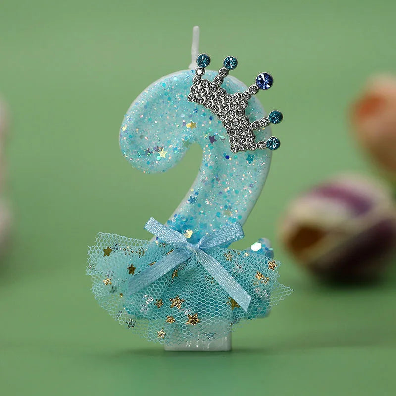 A light blue glitter candle shaped like the number 2 with a silver crown decoration and a sheer blue tulle skirt.
