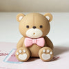 A light brown teddy bear figurine with pink accents and paw prints sitting on a white surface.