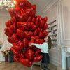 A woman stands next to a large arrangement of red heart-shaped foil balloons in a brightly lit room.