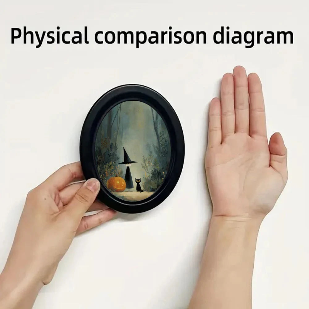 A hand holds an oval framed piece of wall art featuring a witch, a black cat, and woodland scenery.