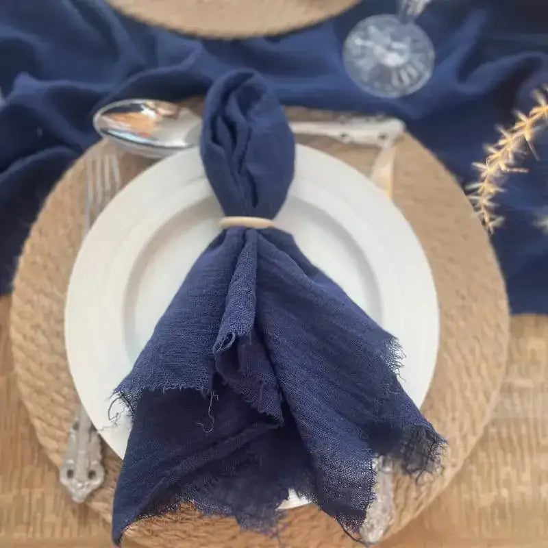 A dark navy blue fringed cotton napkin tied in a bow resting on a white plate and woven mat.