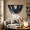 A bohemian macrame wall tapestry featuring deep blue knotted fibers hangs on a beige wall above a light gray sofa.