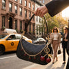 A hand holds a black leather jewelry bag with gold chain and red cherry charms in an urban street setting.