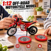 A red 1:12 scale off-road motorcycle model is shown with spare wheels and accessories on a wooden table.