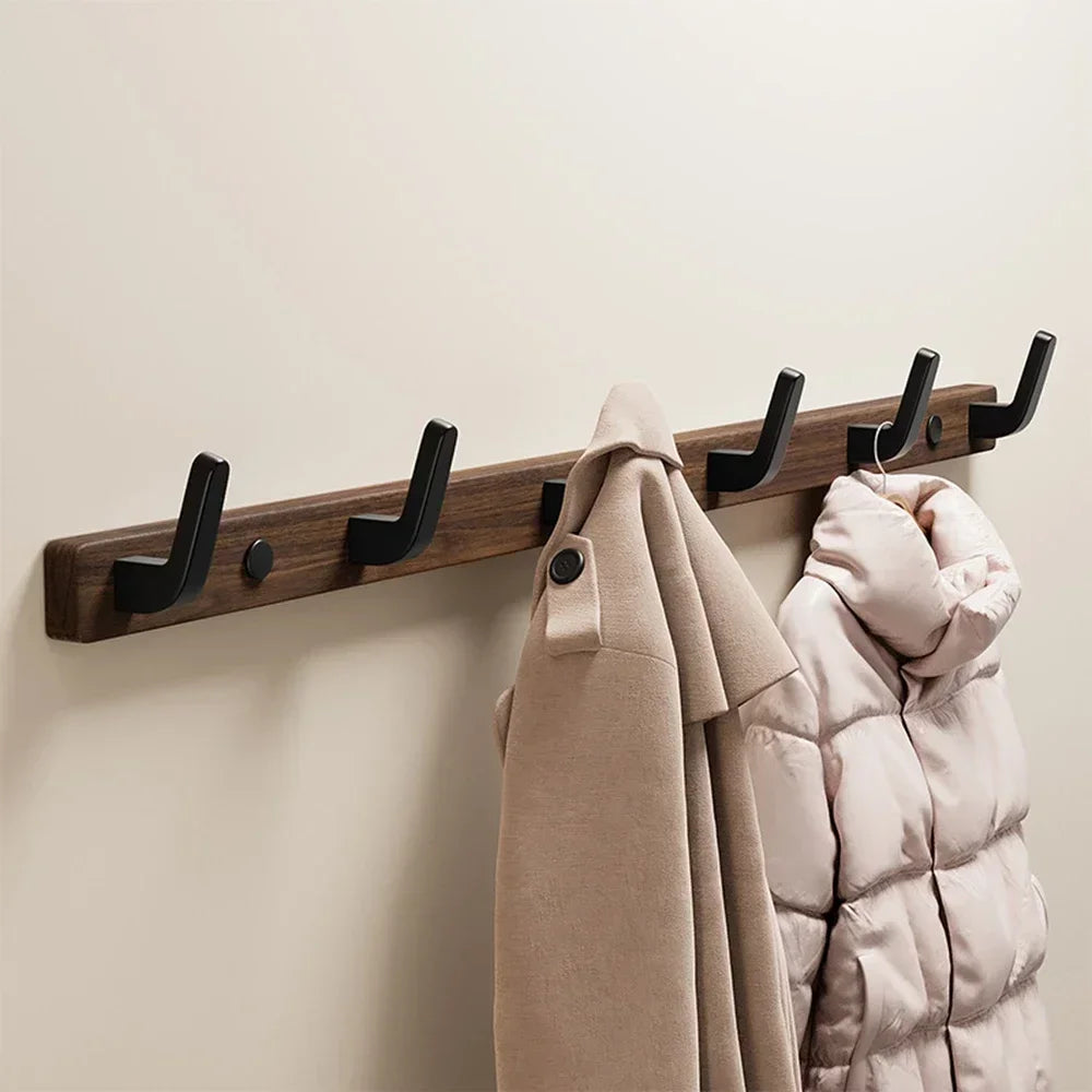 A walnut wood coat rack with eight hooks and a storage shelf mounted on a wall, holding a tan coat and a pink quilted jacket.