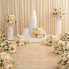 A white tiered cake display surrounded by cream and blush roses and hydrangeas on a wooden table.
