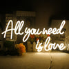 A glowing neon sign spelling out All you need is love illuminates a bouquet of flowers in a dark setting.