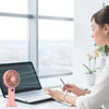 A woman works at a desk with a laptop and a pink portable rechargeable handheld fan on the table.