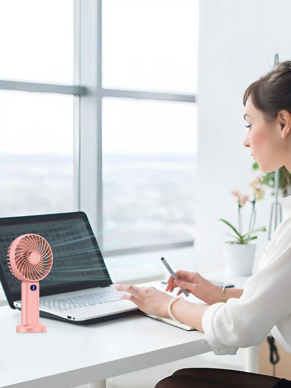 A woman works at a desk with a laptop and a pink portable rechargeable handheld fan on the table.