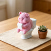 Figurine de porc en céramique rose assise sur une cuvette blanche, à côté d'une petite plante succulente.