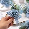 A hand holds a dusty blue silk rose boutonniere featuring soft blue and green foliage.