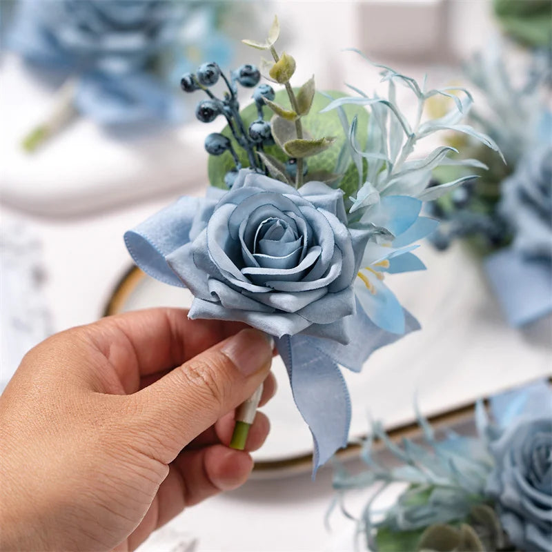 A hand holds a dusty blue silk rose boutonniere featuring soft blue and green foliage.