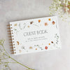 A white personalized wedding guest book with gold-toned spiral binding and floral watercolor accents on a light gray backgrou