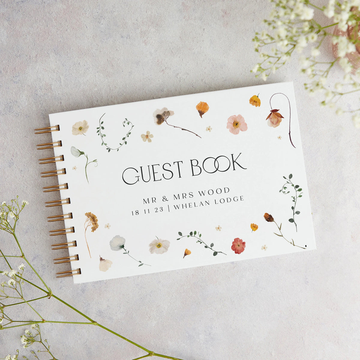A white personalized wedding guest book with gold-toned spiral binding and floral watercolor accents on a light gray backgrou