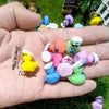 A hand holds a set of mini resin cruise ducks with sunglasses in assorted colors on green grass.