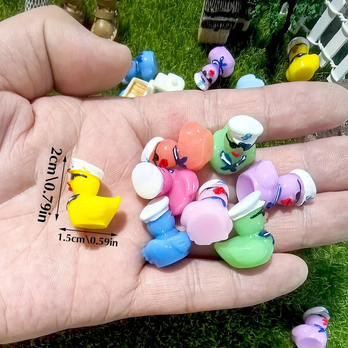A hand holds a set of mini resin cruise ducks with sunglasses in assorted colors on green grass.