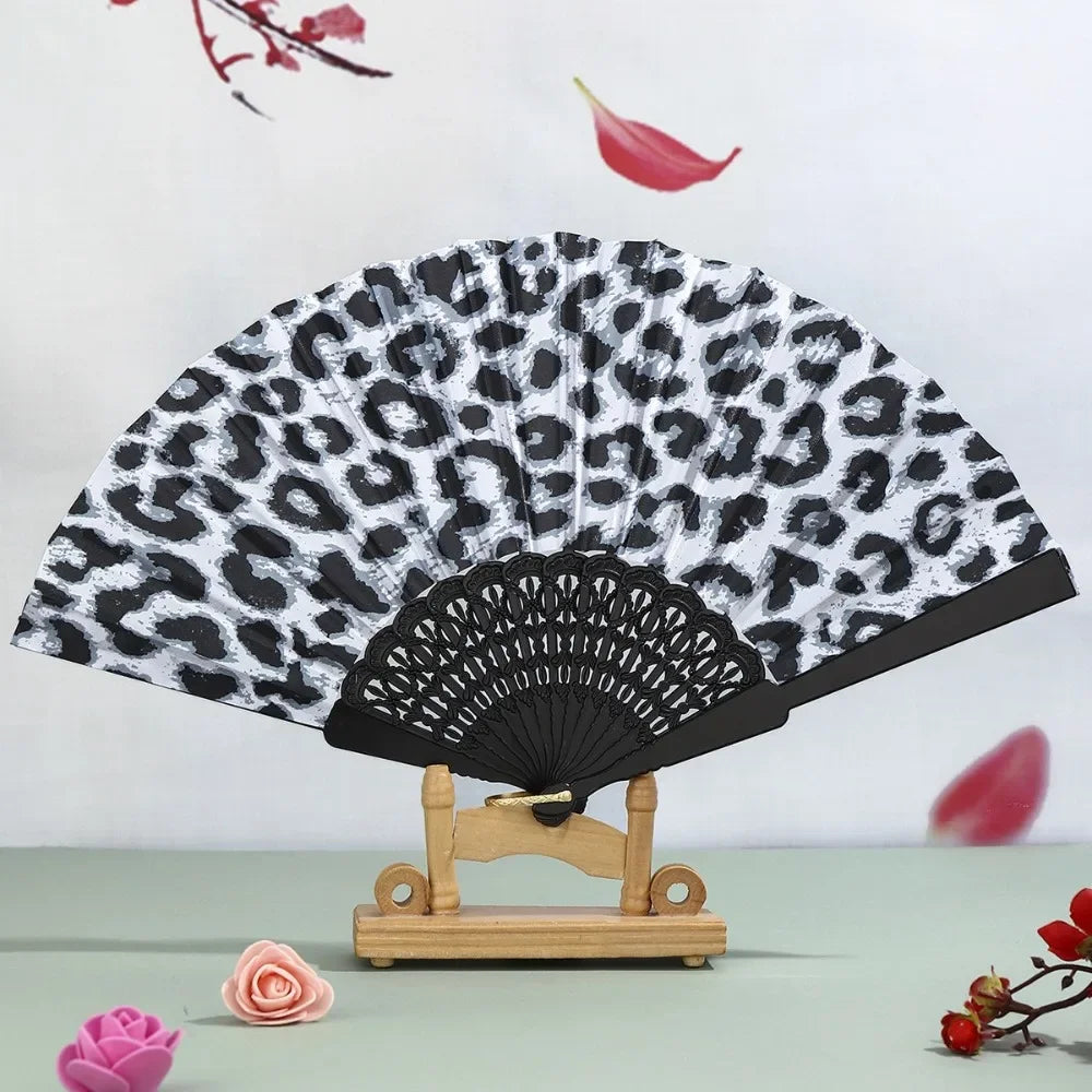 A black and white leopard print folding hand fan with a black handle displayed on a wooden stand.