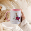 A hand holds a pink ceramic coffee mug with a gold letter initial decorated with floral elements.