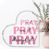A clear acrylic heart-shaped sign with the text Pray on it pray over it pray through it displayed next to some greenery.