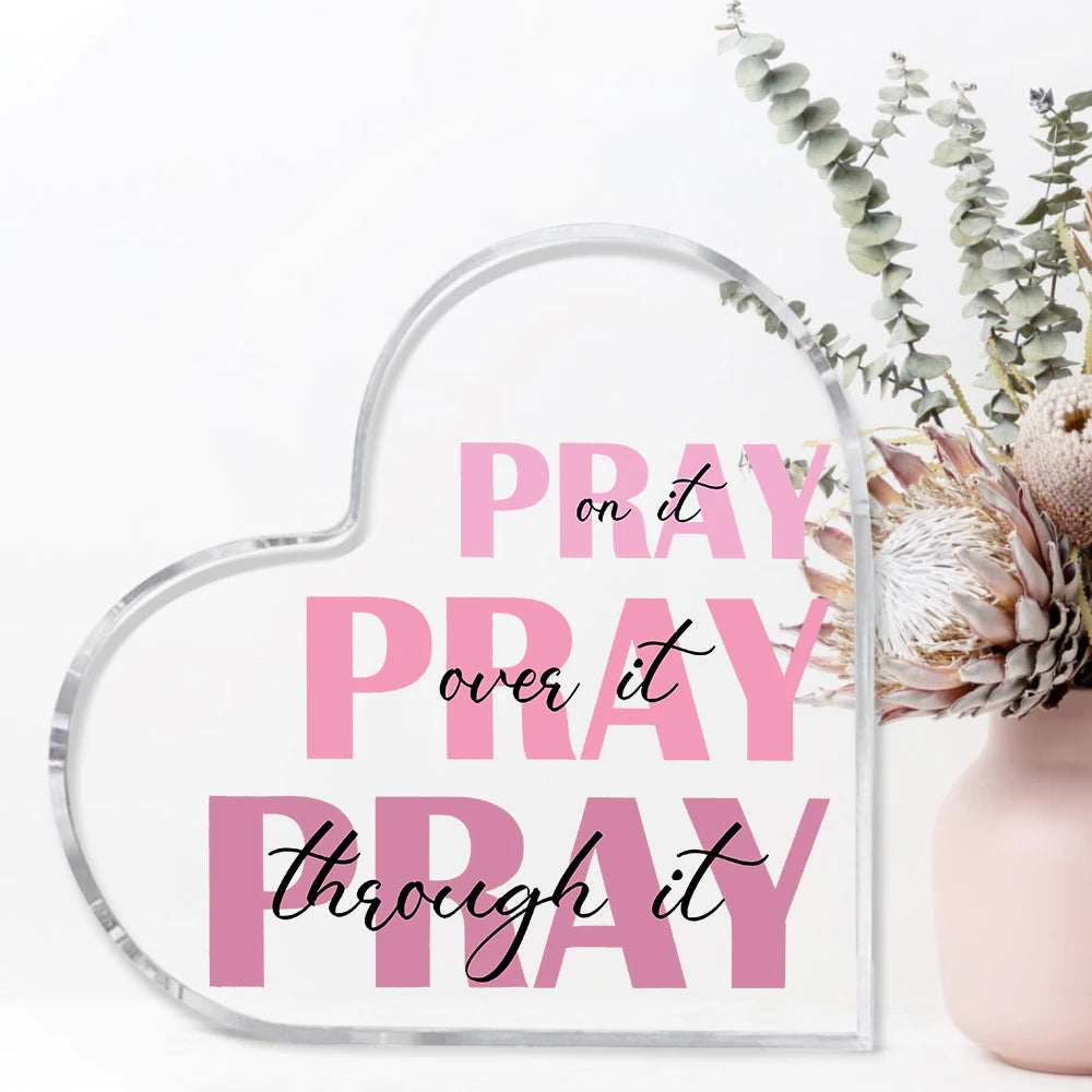 A clear acrylic heart-shaped sign with the text Pray on it pray over it pray through it displayed next to some greenery.