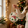 A hand holds a finished Christmas ornament made of pearl and diamond painting on a string near a decorated Christmas tree.