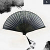A black vintage silk bamboo folding hand fan is shown open, displaying delicate, light-colored floral patterns against a whit