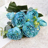 A bouquet of artificial blue silk roses and peonies with green leaves and small accents on a white surface.