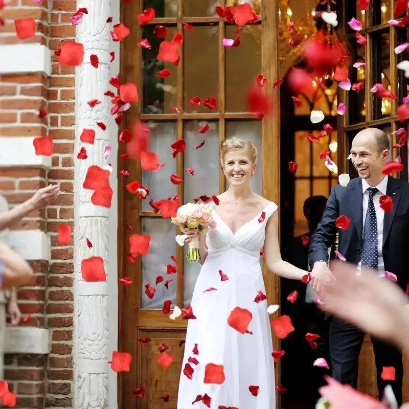 A smiling bride in a white dress is showered with red rose petals indoors.