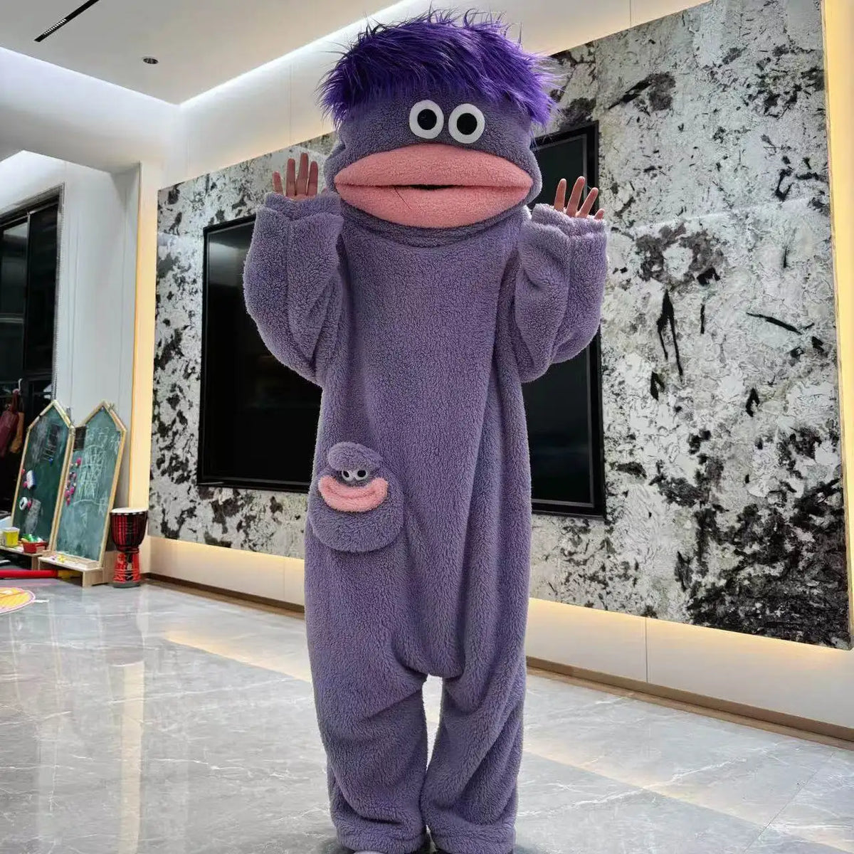 A purple, fuzzy monster coral fleece hooded sleepsuit with large eyes and a pink mouth standing in a modern room.