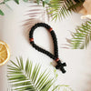 A black prayer rope with wooden beads and a cross charm is displayed on a white background surrounded by green leaves.
