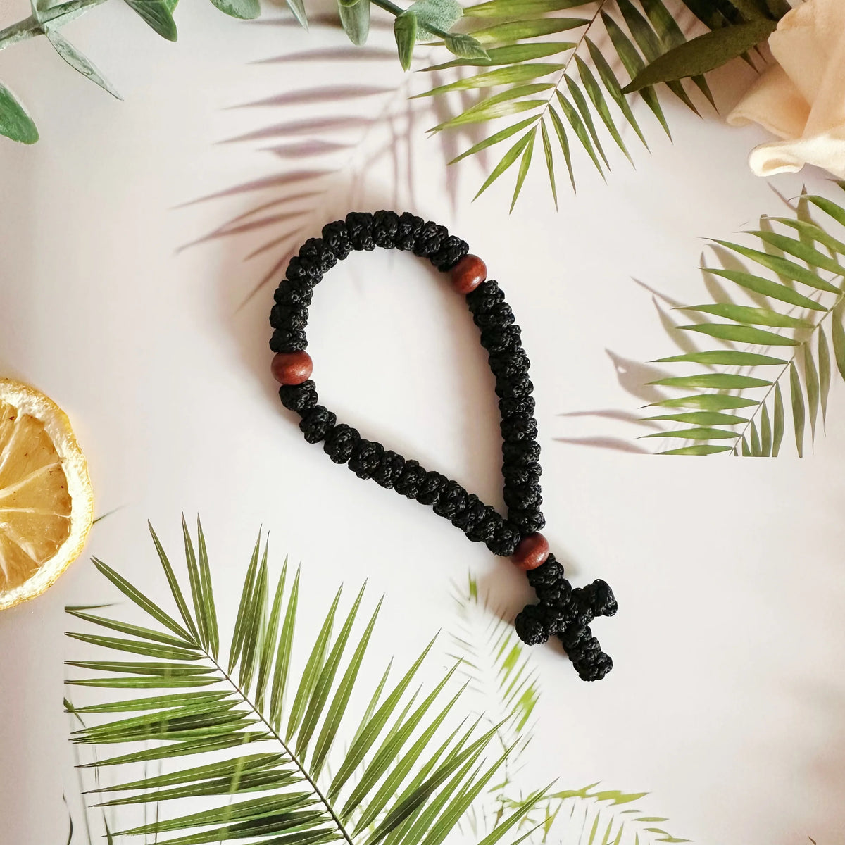 A black prayer rope with wooden beads and a cross charm is displayed on a white background surrounded by green leaves.