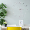 A modern silver acrylic wall clock with twelve hour markers is mounted on a light gray wall above a wooden table.