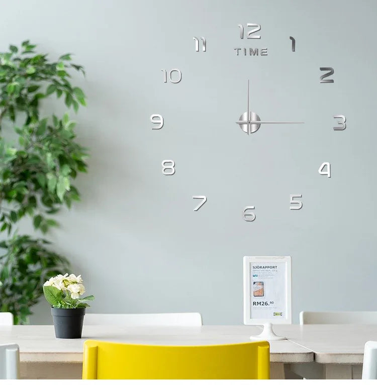 A modern silver acrylic wall clock with twelve hour markers is mounted on a light gray wall above a wooden table.
