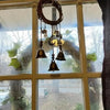 A rustic metal wreath with hanging bells is displayed in a window frame surrounded by green foliage.