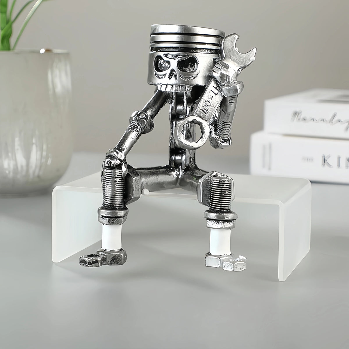 A silver mechanical piston skull sculpture is displayed on a white rectangular stand.
