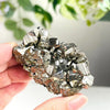 A hand holds a cluster of sparkling metallic pyrite crystals against a blurred green background.