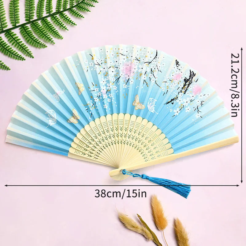A light blue bamboo folding hand fan with floral accents and a blue tassel displayed on a pink background.