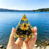 Hand holding a clear resin pyramid with natural obsidian and a gold-colored coil against a blue lake background